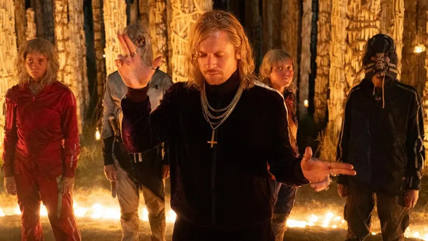 A blond man in black with a bone necklace standing before robed figures in a fire-lit forest